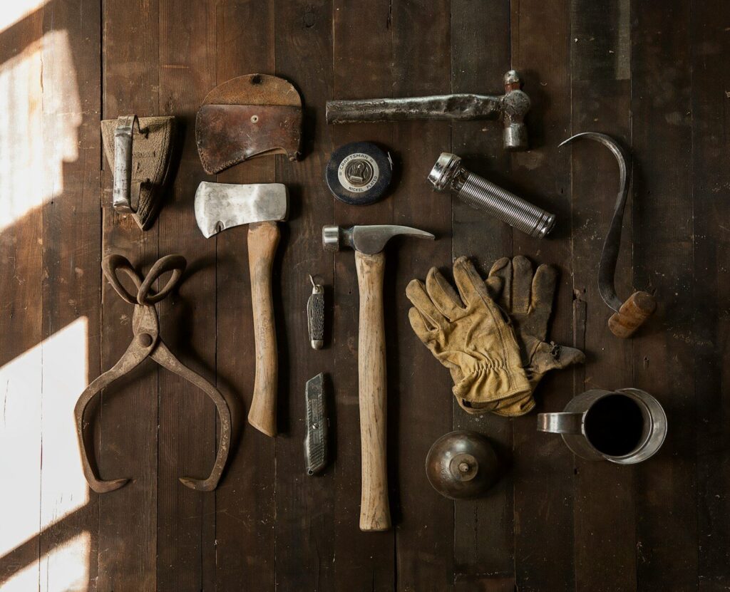 Vintage tools including an axe, hammer, gloves, and metal implements on dark wooden planks